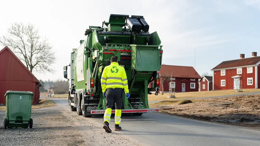 En sopchaufför tömmer kärl utanför röda stugor med vita knutar.